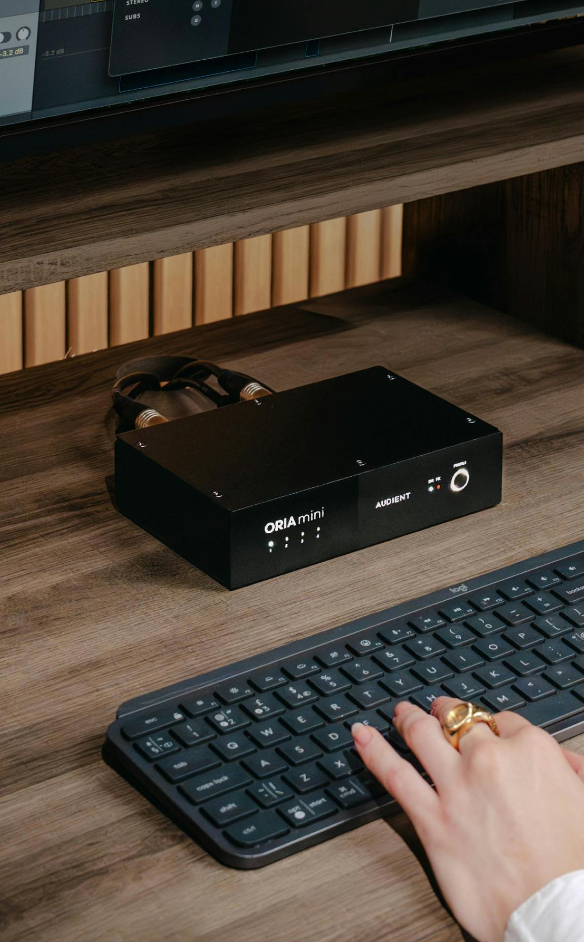 Close-up of Audient ORIA Mini audio interface on wooden studio desk with cables connected, placed above keyboard while user types.