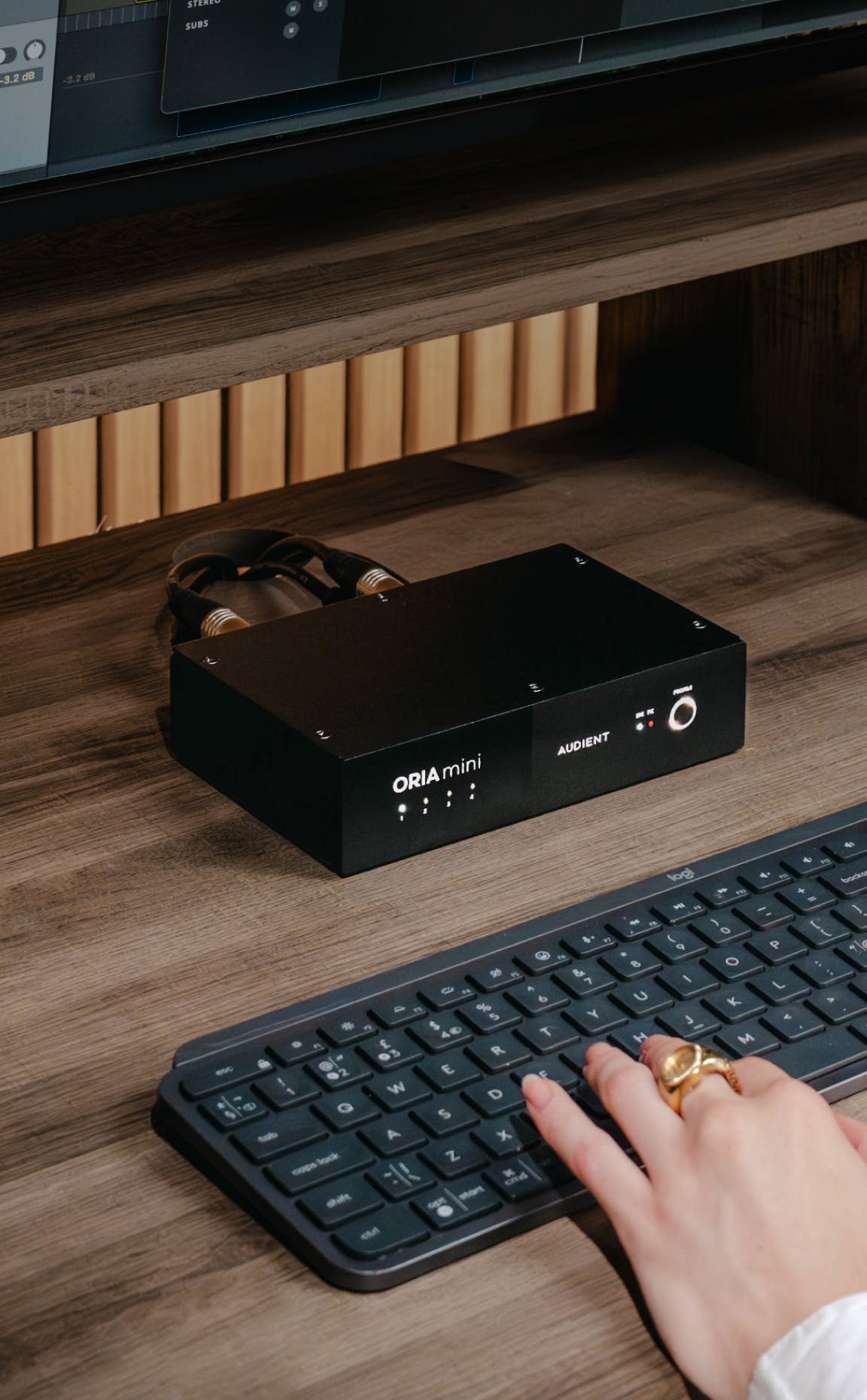 Close-up of Audient ORIA Mini audio interface on wooden studio desk with cables connected, placed above keyboard while user types.
