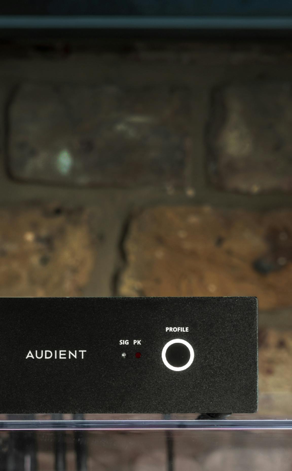 Close-up of Audient ORIA Mini front panel with illuminated profile knob and signal indicators, placed against exposed brick wall background.