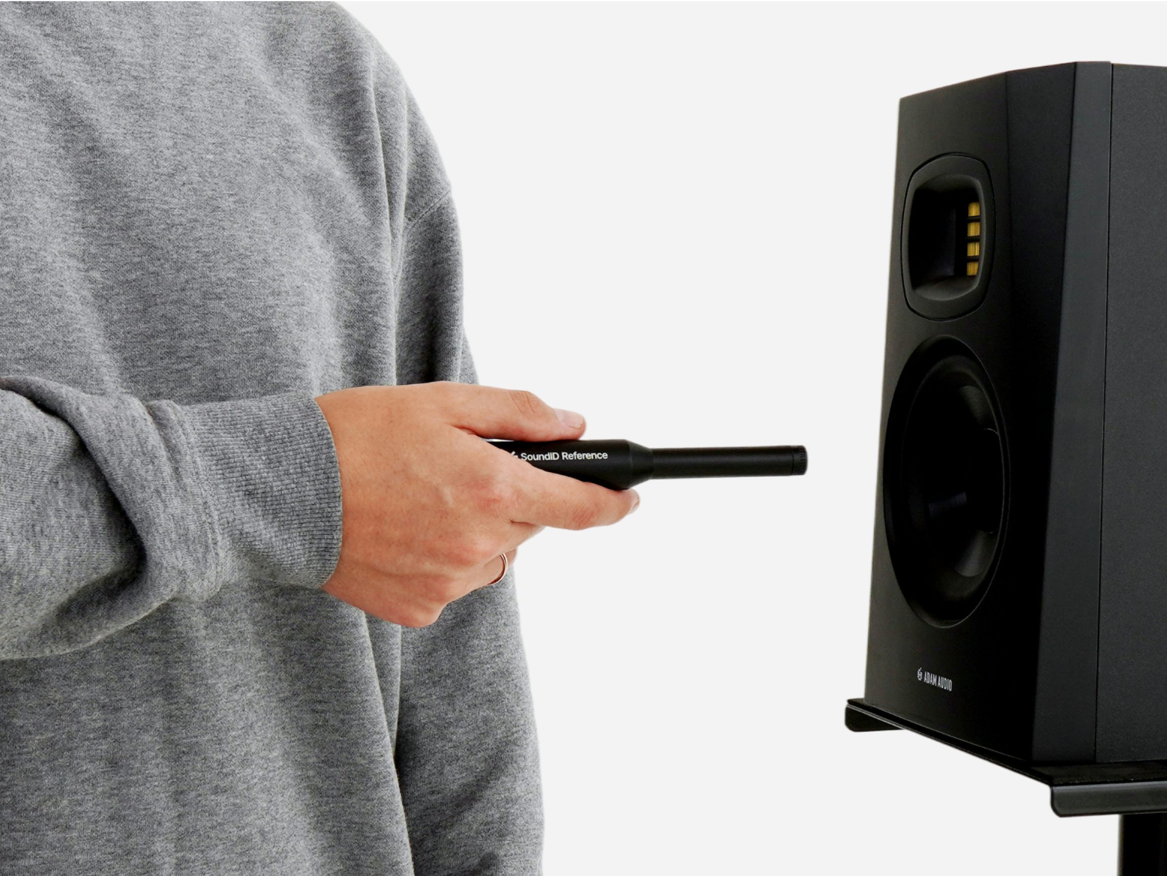 Hand holding SoundID Reference measurement microphone aimed at black ADAM Audio studio monitor on stand, grey sweatshirt.